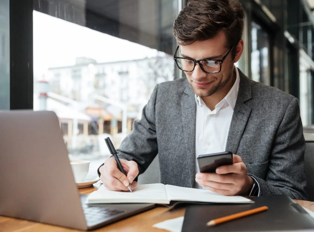 smiling-businessman-eyeglasses-sitting-by-table-cafe-with-laptop-computer-while-using-smartphone-writing-something